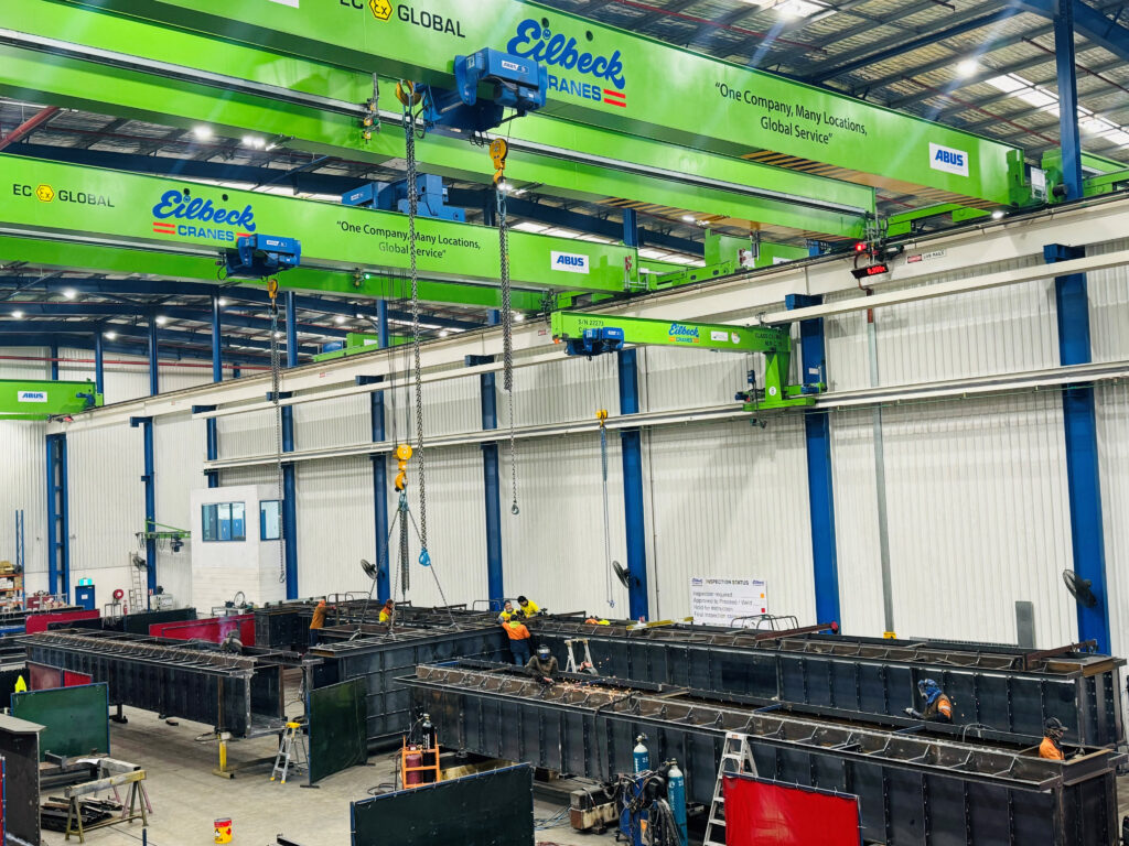 Welders work on large steel weldments in the Eilbeck fabrication bay under green overhead cranes