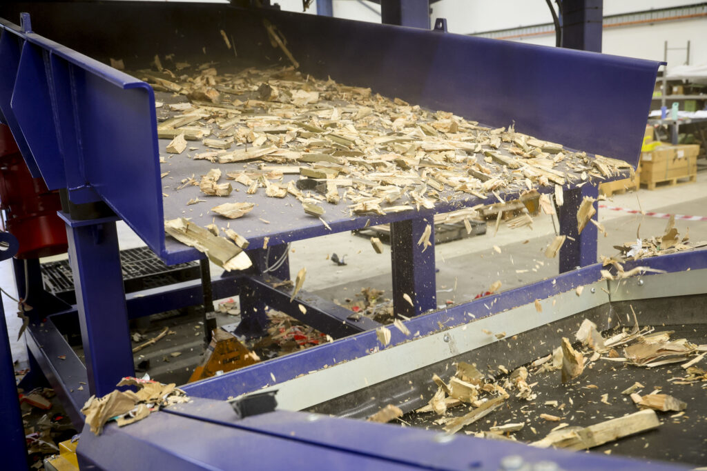 Shredded timber falling from a blue vibration table onto a belt conveyor
