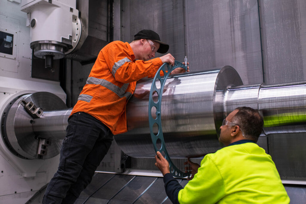 Two Eilbeck machinists measure a large machined shaft with an outside micrometer